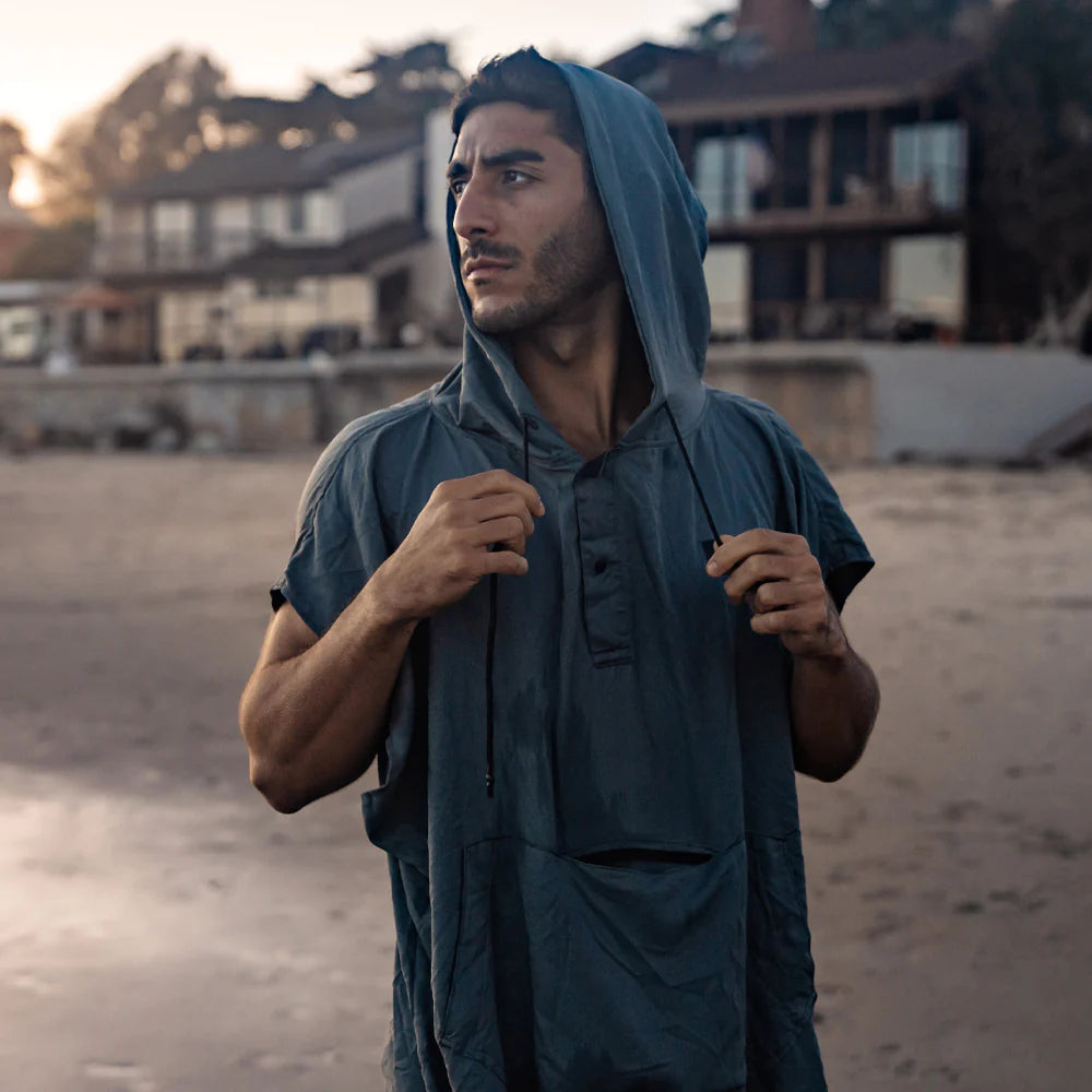 An image of a person standing on a beach at sunset, wearing a blue hooded Mataponcho. The individual is adjusting the hood drawstrings with both hands, with houses and trees visible in the blurred background.