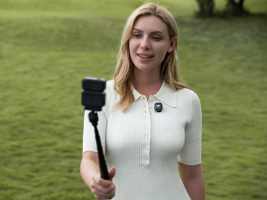 Woman holding a selfie stick with a camera attachment in a grassy outdoor setting