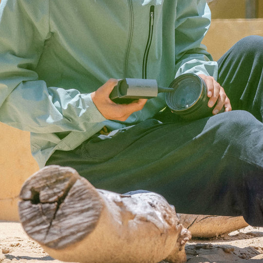 Person in light green jacket and dark pants sitting on a log, holding a VSGO Pocket Beast Blower.