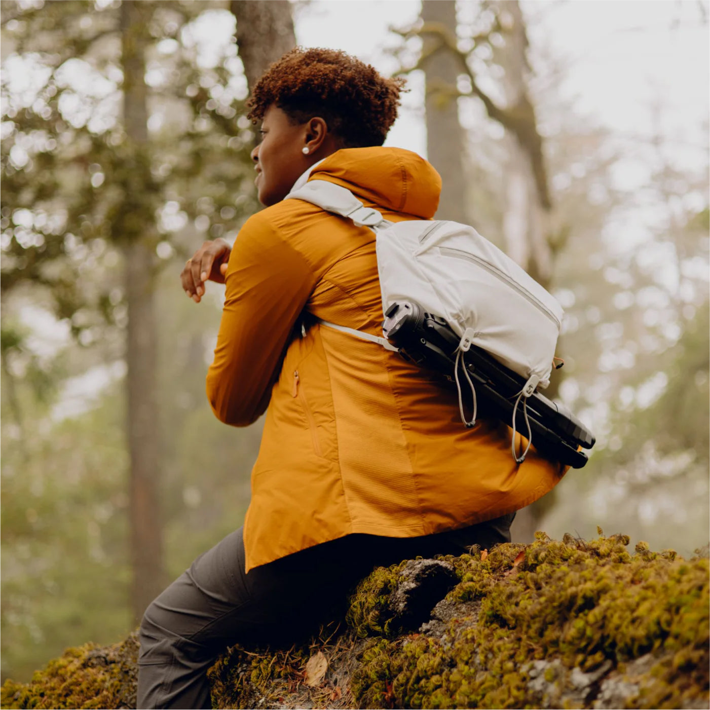 a person with orange jacket sitting on a rock wearing Peak Design Sling white infront of a forest scenery.