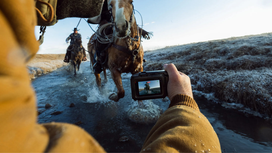 Person taking a photo of two horses crossing a stream with a camera held up to the lens.