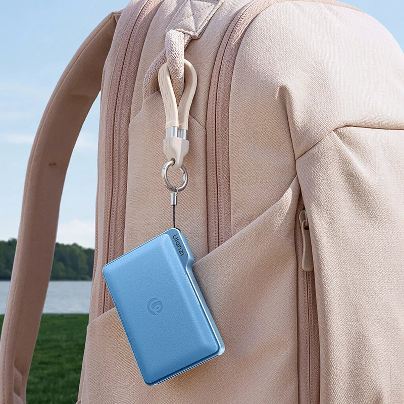 A light blue rectangular card holder or wallet attached to a beige backpack with a white clip and key ring, photographed outdoors near water and grass.