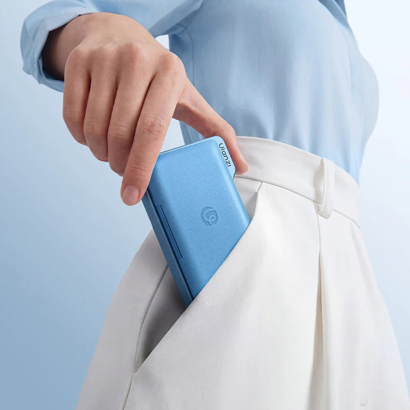 A close-up of a person's hand sliding a light blue rectangular electronic device into the pocket of white trousers, with a light blue shirt visible.