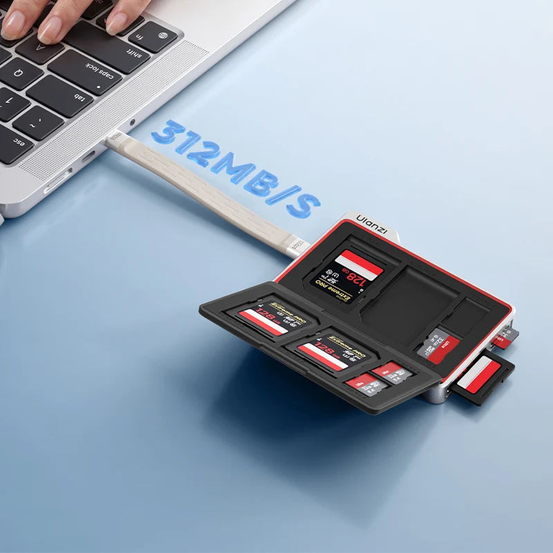 A compact black and red multi-card SD card reader and memory card hub with multiple cards inserted, shown plugged into a silver laptop on a light blue surface. A hand is visible at the top left, and blue text reads '312MB/S'.