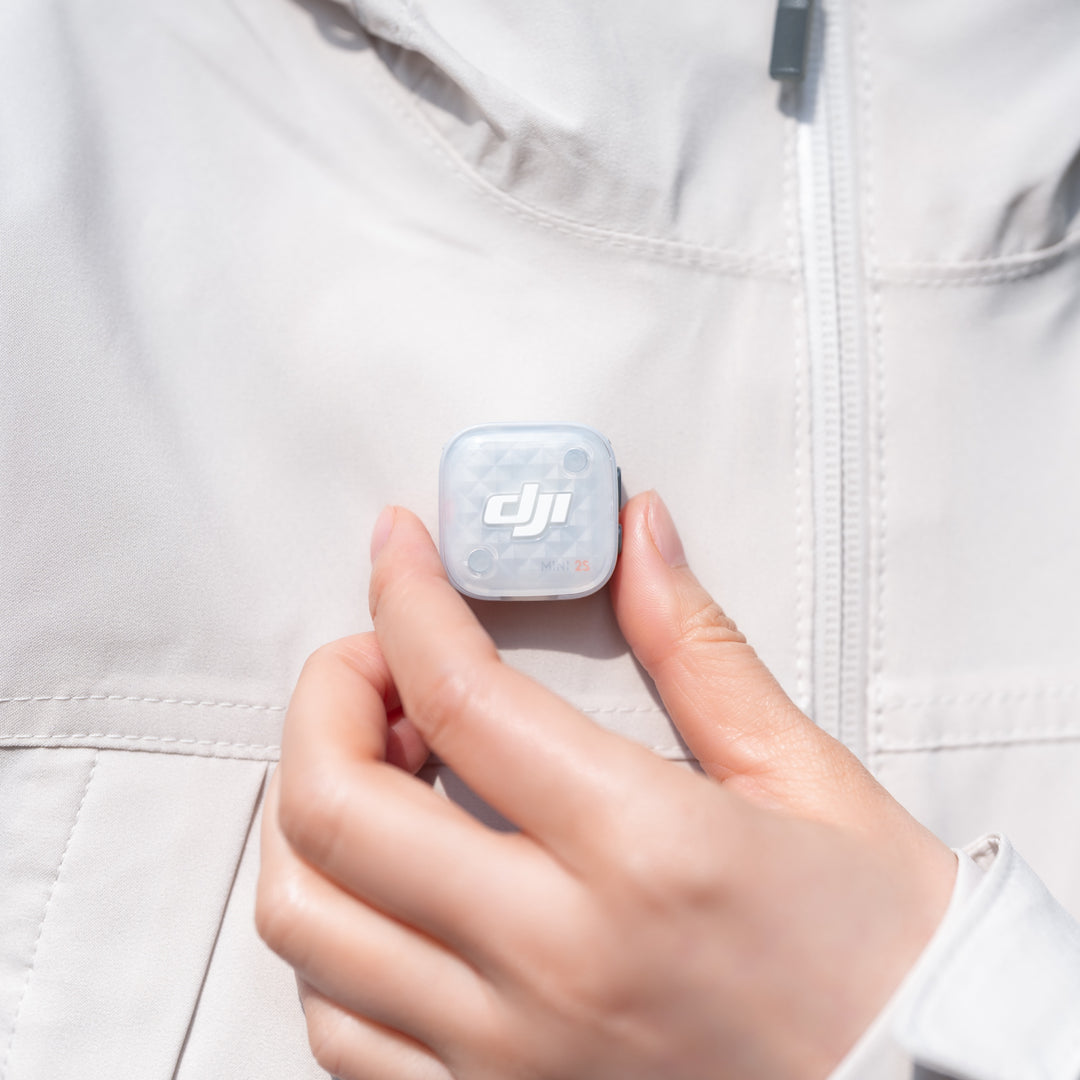 A close-up of a hand holding a DJI mic mini 2s transmitter against a light-colored zippered jacket.