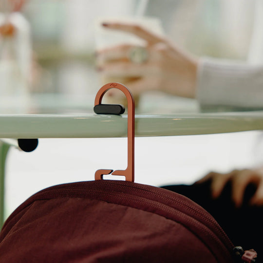A copper-colored metal backpack clip hanging from a glass table, holding a burgundy backpack. A person&