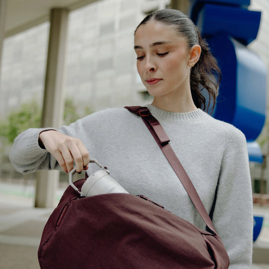 A woman in a light gray knit sweater carries a large burgundy shoulder bag while pouring water from a silver bottle into it. The setting is outdoors with a modern building and blue sculpture in the background.