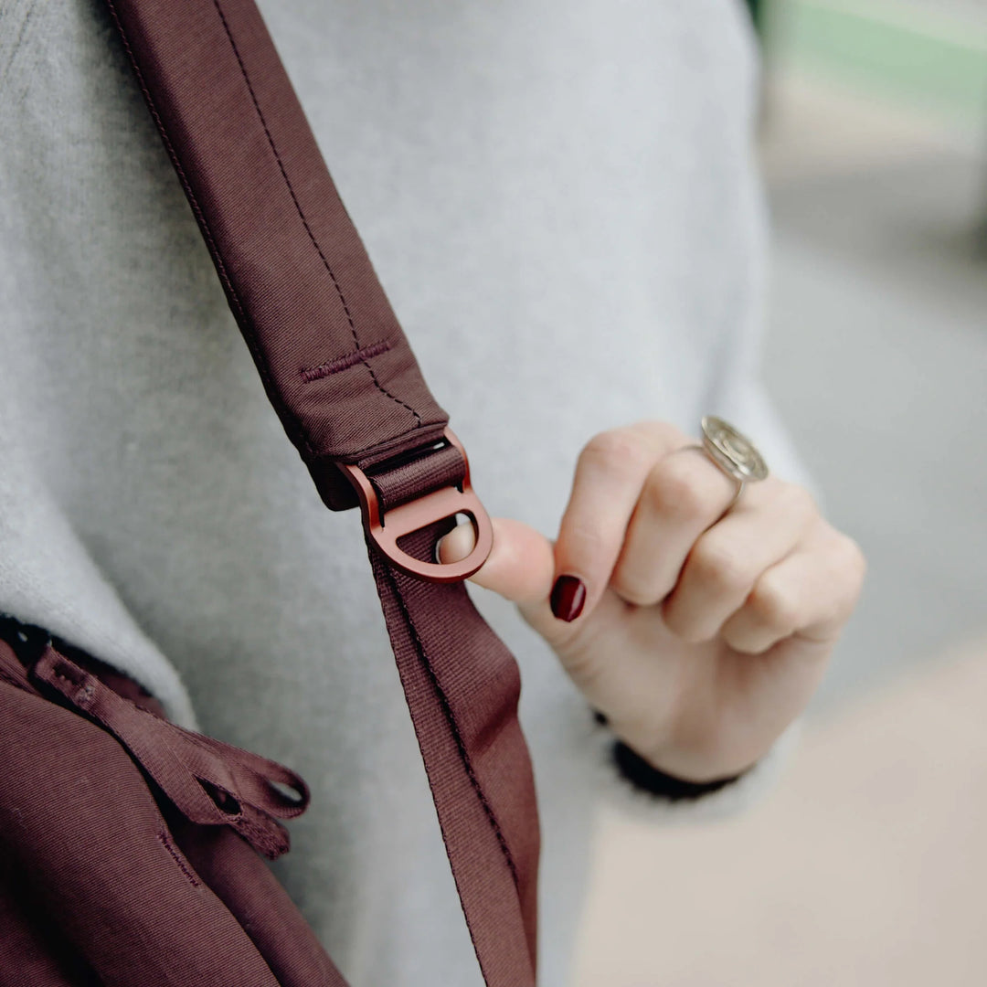 Close-up of a person wearing a gray sweater and adjusting the strap of a dark brown crossbody bag with a rose gold metal buckle.