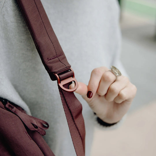 Close-up of a person wearing a gray sweater and adjusting the strap of a dark brown crossbody bag with a rose gold metal buckle.