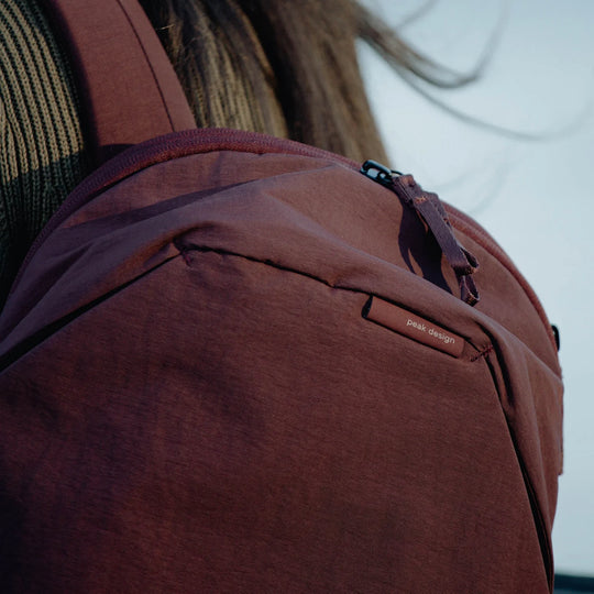 Close-up of a person wearing a burgundy Peak Design backpack, showing the brand label on the front pocket and the textured fabric under warm natural light.