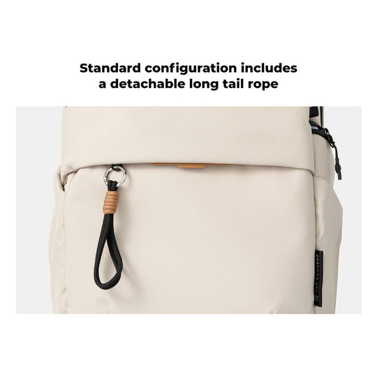 Close-up of a light beige backpack with a detachable black tail rope hanging from a front zipper, with text above reading &
