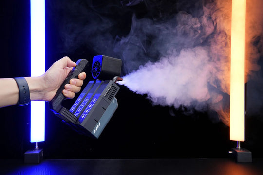 Hand holding a PMI SmokeNINJA PRO - Handheld Fog Machine emitting smoke against a dark background with colorful lights.