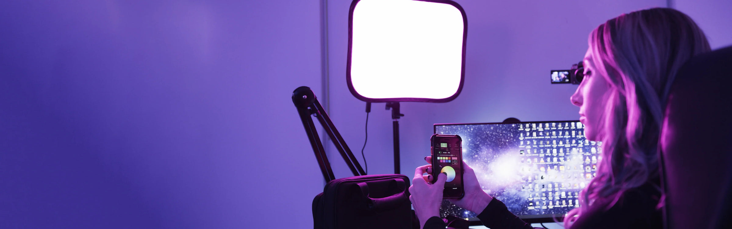Person using a smartphone with a light panel and computer screen in a dimly lit room.