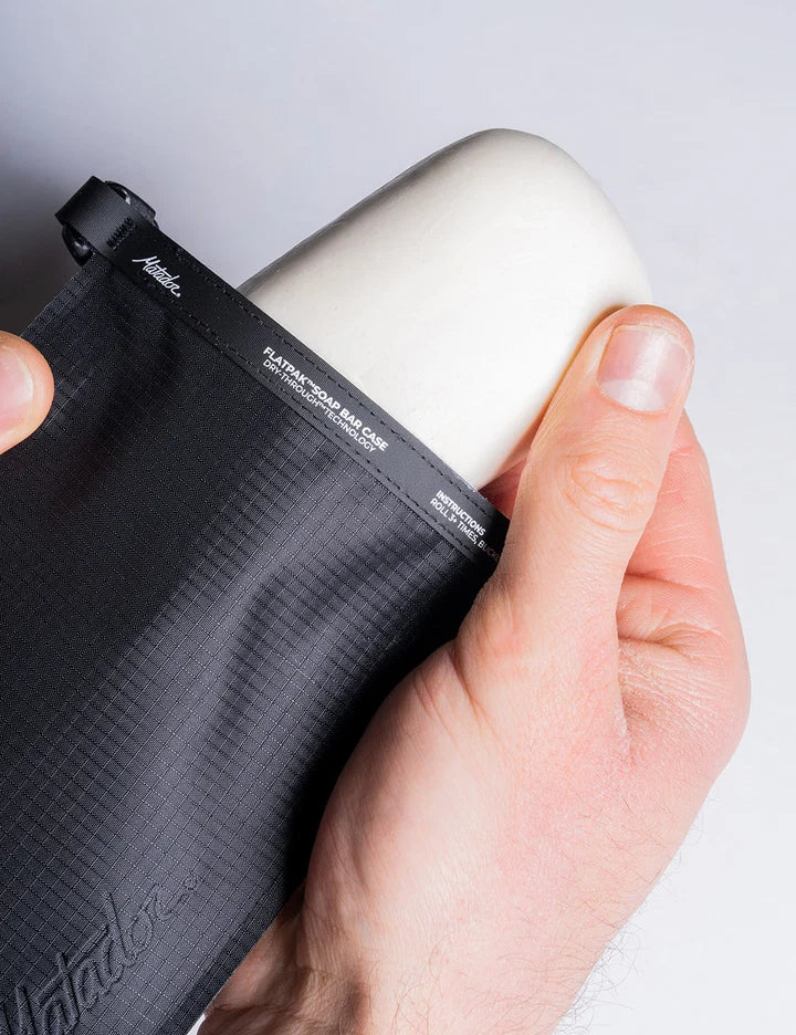A close-up of a hand holding a black waterproof bag with a white cylindrical insert or cap, showing the brand name 'Klatu' printed on the black strap.