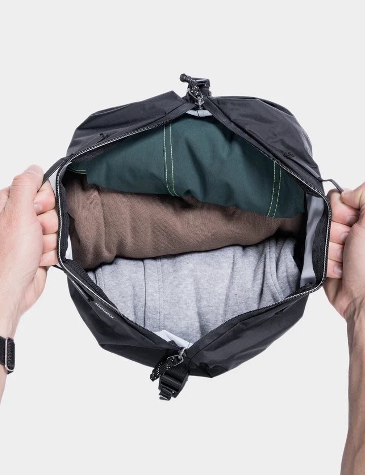 A black waterproof packing cube or dry bag being held open by two hands, showing folded clothes inside in shades of green, brown, and gray, against a light gray background.