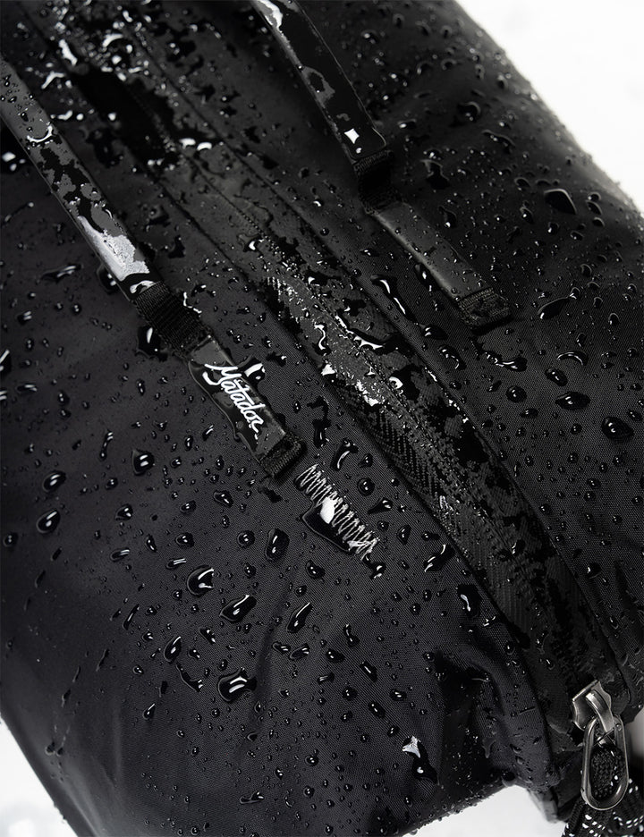 Close-up of a black waterproof bag covered in water droplets, showing a zipper and a small white script logo tag.