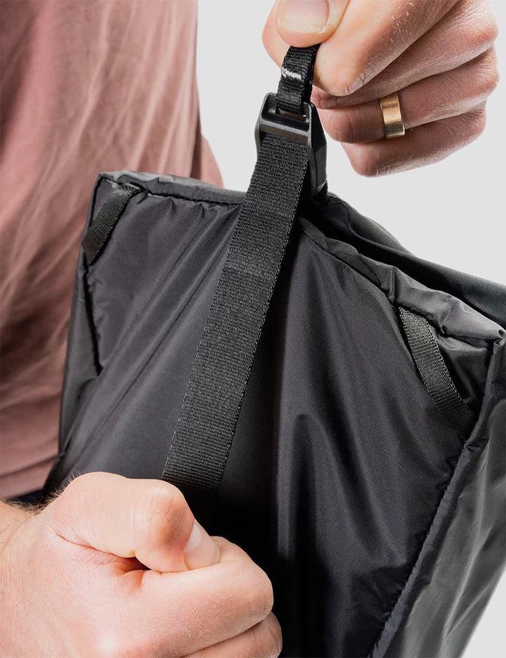 Close-up of a person's hands adjusting a black adjustable webbing strap on a black puffer bag, with a gold ring visible on one finger.