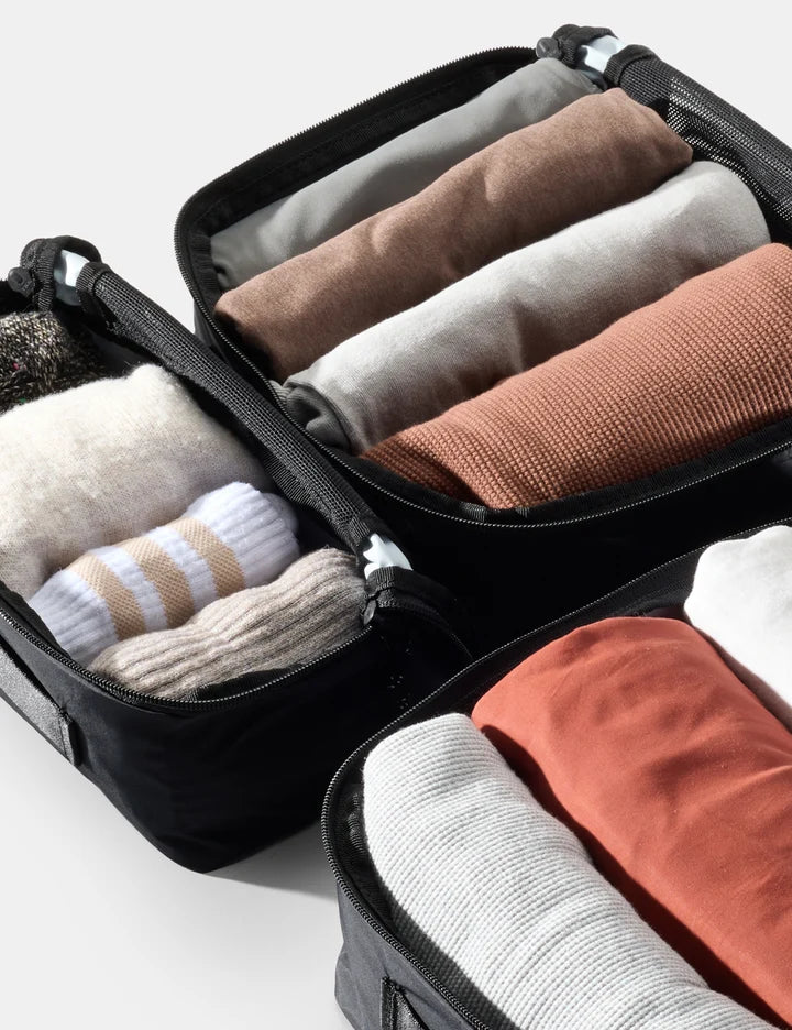 Three black zippered packing cubes filled with neatly folded travel clothes and socks, arranged on a light gray background.