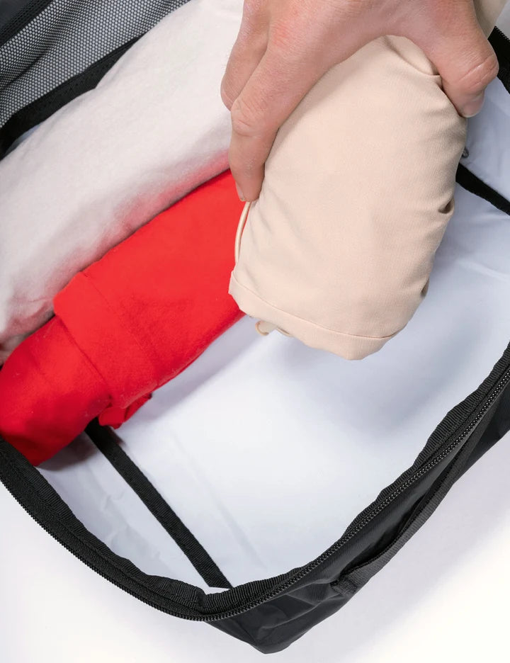 A close-up of a hand placing a beige folded garment into a black zippered packing cube, alongside a red folded item and a white inner liner.