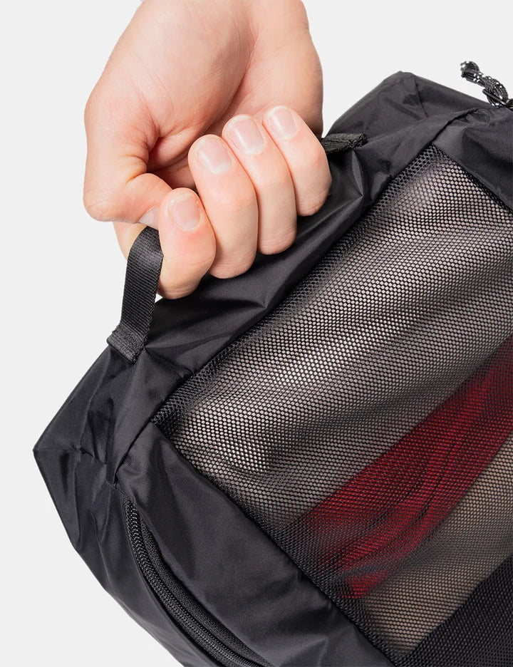 A close-up of a hand holding the zipper pull of a black mesh travel packing cube, showing the texture of the fabric and a glimpse of red packing material inside, set against a plain light gray background.
