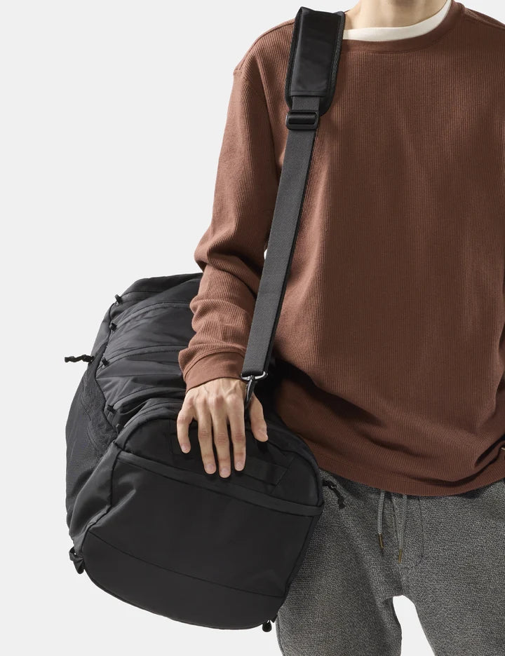 A person wearing a brown sweatshirt and gray sweatpants carries a large black duffel bag with a wide shoulder strap against a light gray background.