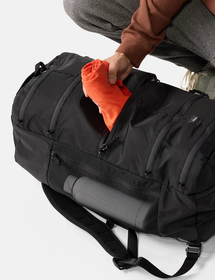 A black duffel bag with multiple zippered compartments and a shoulder strap, shown with a person's hand placing an orange towel or blanket into a front pocket. A gray rolled mat is also visible inside the bag against a clean white background.