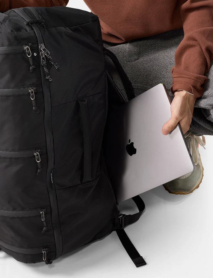 A black backpack shown close up with a person seated beside it, holding a silver laptop inside the backpack. The person wears a rust-brown sweater, gray pants, and tan shoes.
