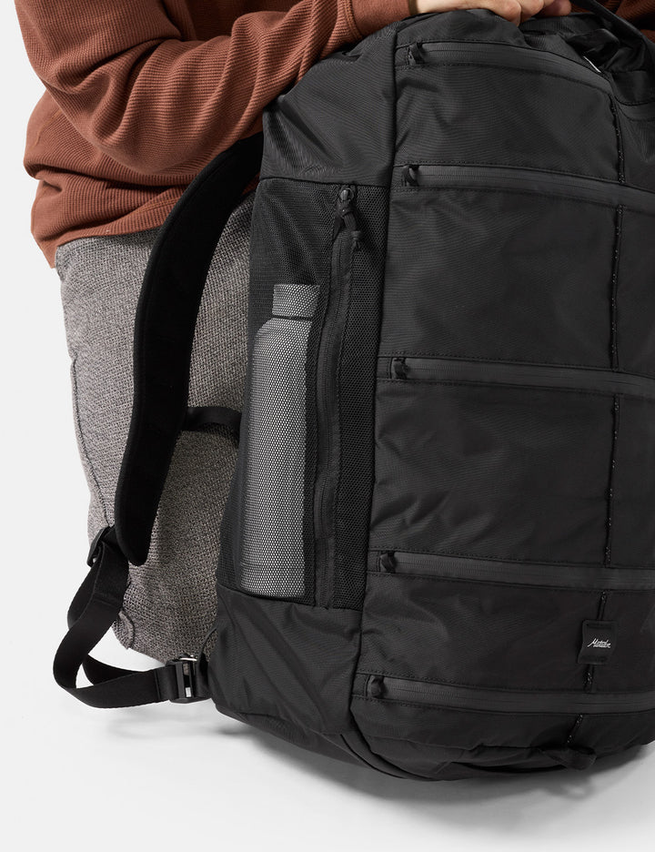 A close-up of a black backpack being worn, with part of a person's torso visible in a brown ribbed top and gray pants. The backpack has multiple zippered compartments, a side mesh pocket with a gray bottle, and a small black logo patch near the bottom right.