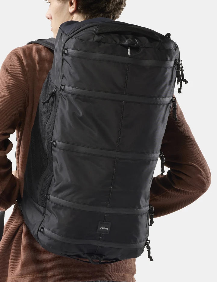 A person wearing a large black technical backpack, shown from the back against a light gray studio background.