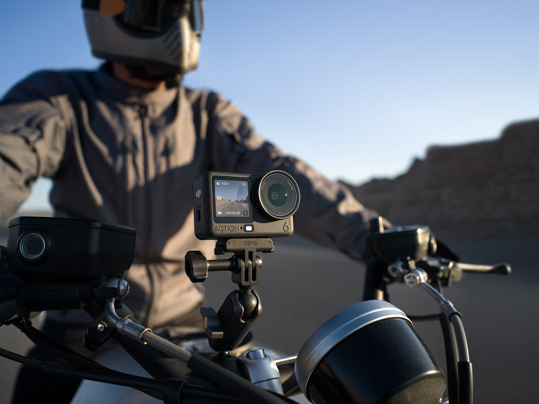 Person riding a motorcycle with a camera mounted on the handlebar, set against a desert landscape.