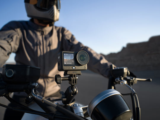 Person riding a motorcycle with a camera mounted on the handlebar, set against a desert landscape.