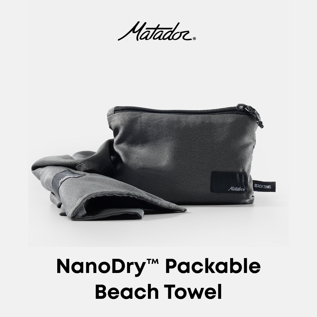 Matador NanoDry Packable Beach Towel set on a light gray background, featuring a dark gray towel and its matching zippered storage pouch with the Matador logo.