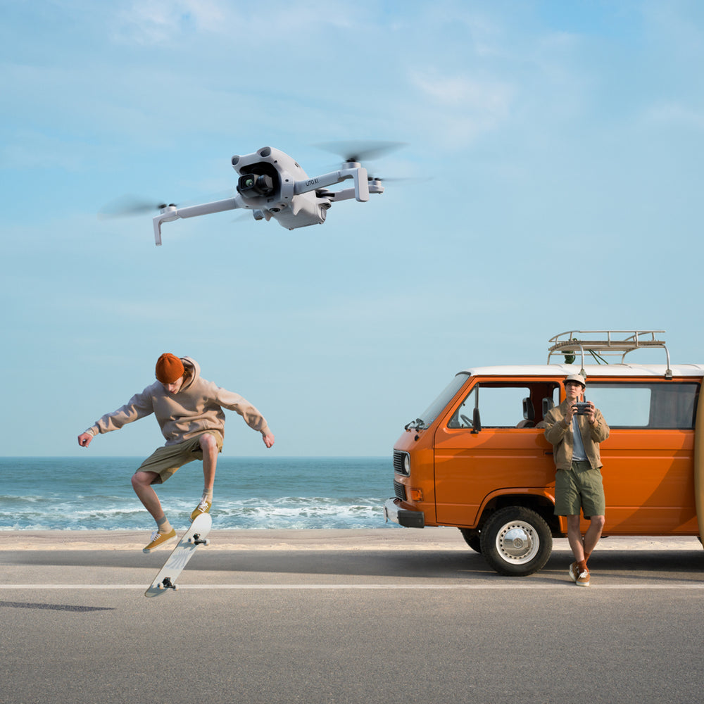 Two people by an orange van with a DJI Lito X1 drone flying above, near the ocean.