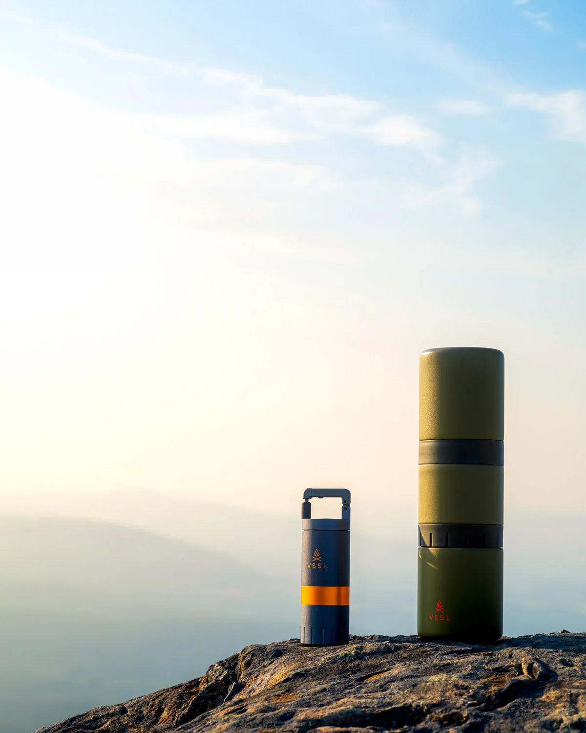 Two cylindrical insulated containers from the brand ZELL, one small blue-and-orange container and one larger olive green container, standing on a rocky mountain peak against a bright sky.