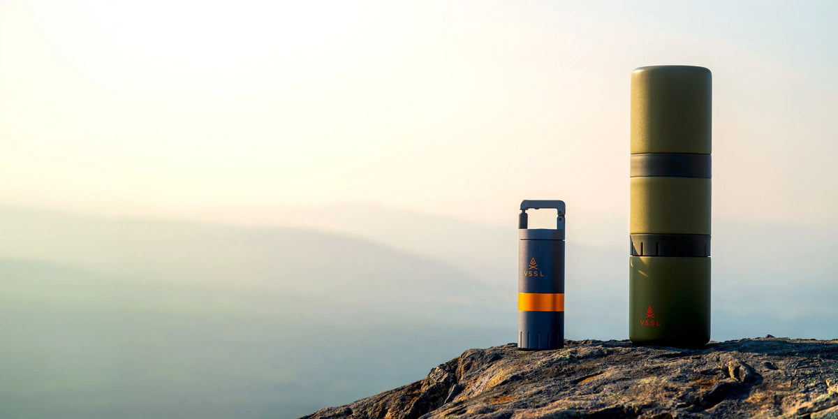 Two cylindrical outdoor trash cans on a rocky mountain peak, with one canister standing upright and the other tipped on its side to show its rectangular opening, against a misty, hazy background.
