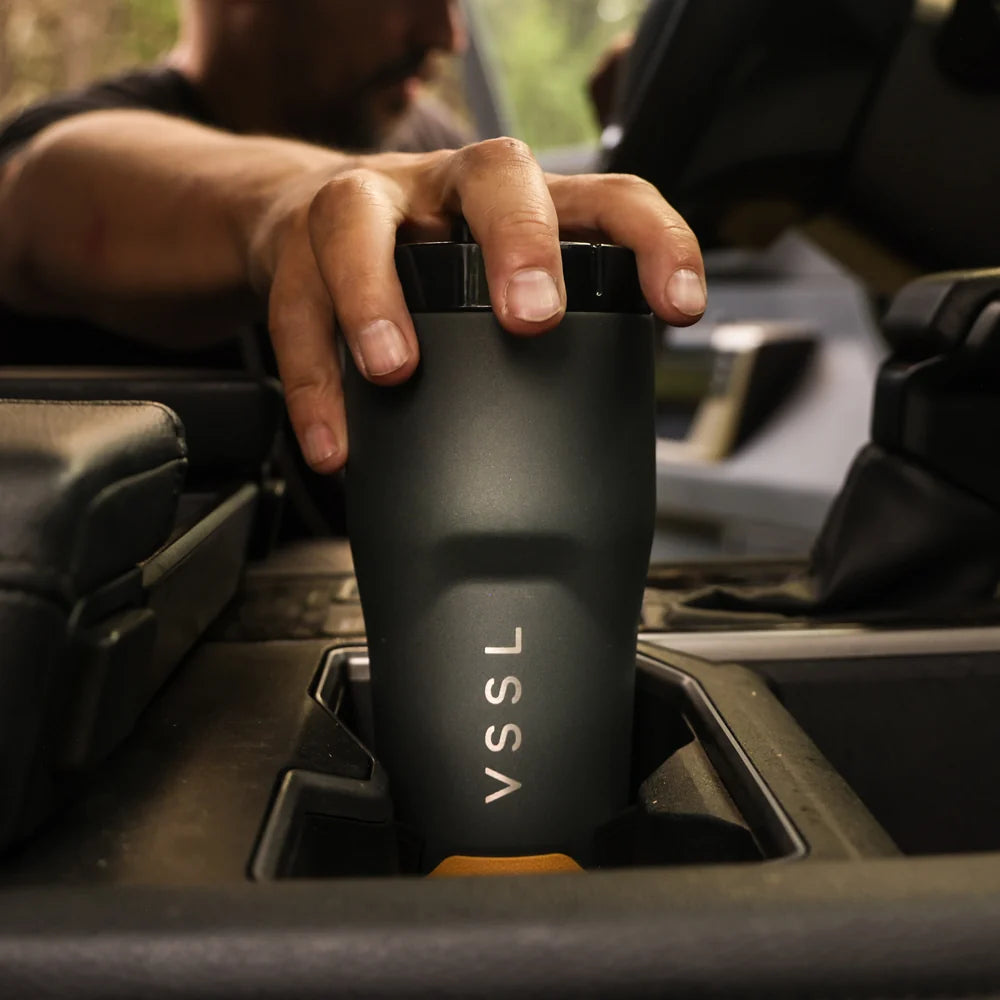 A close-up of a person's hand holding a matte black VSSL insulated tumbler cup inside a car cup holder.
