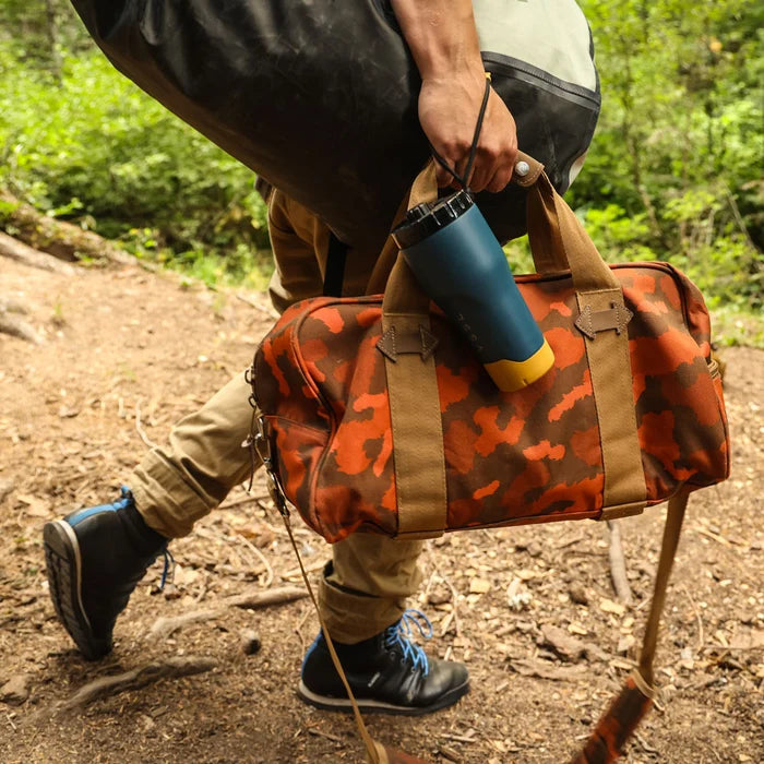 Person carrying a camouflage duffel bag with a blue and yellow container on a trail.