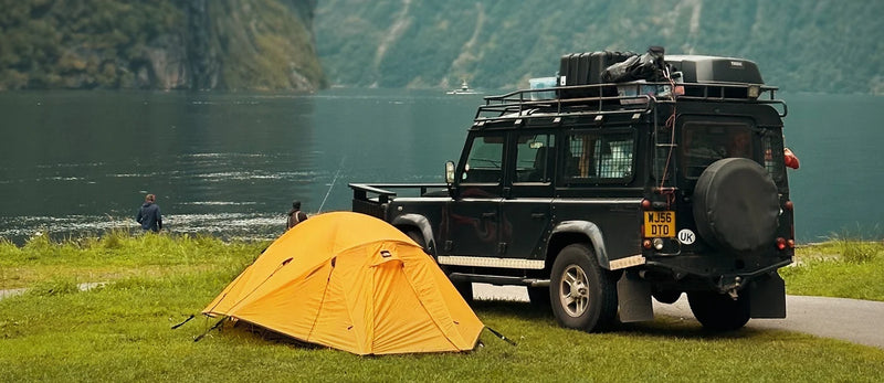 A bright yellow dome camping tent pitched on grass beside a black off-road SUV with roof gear, set by a lake with mountains in the background.