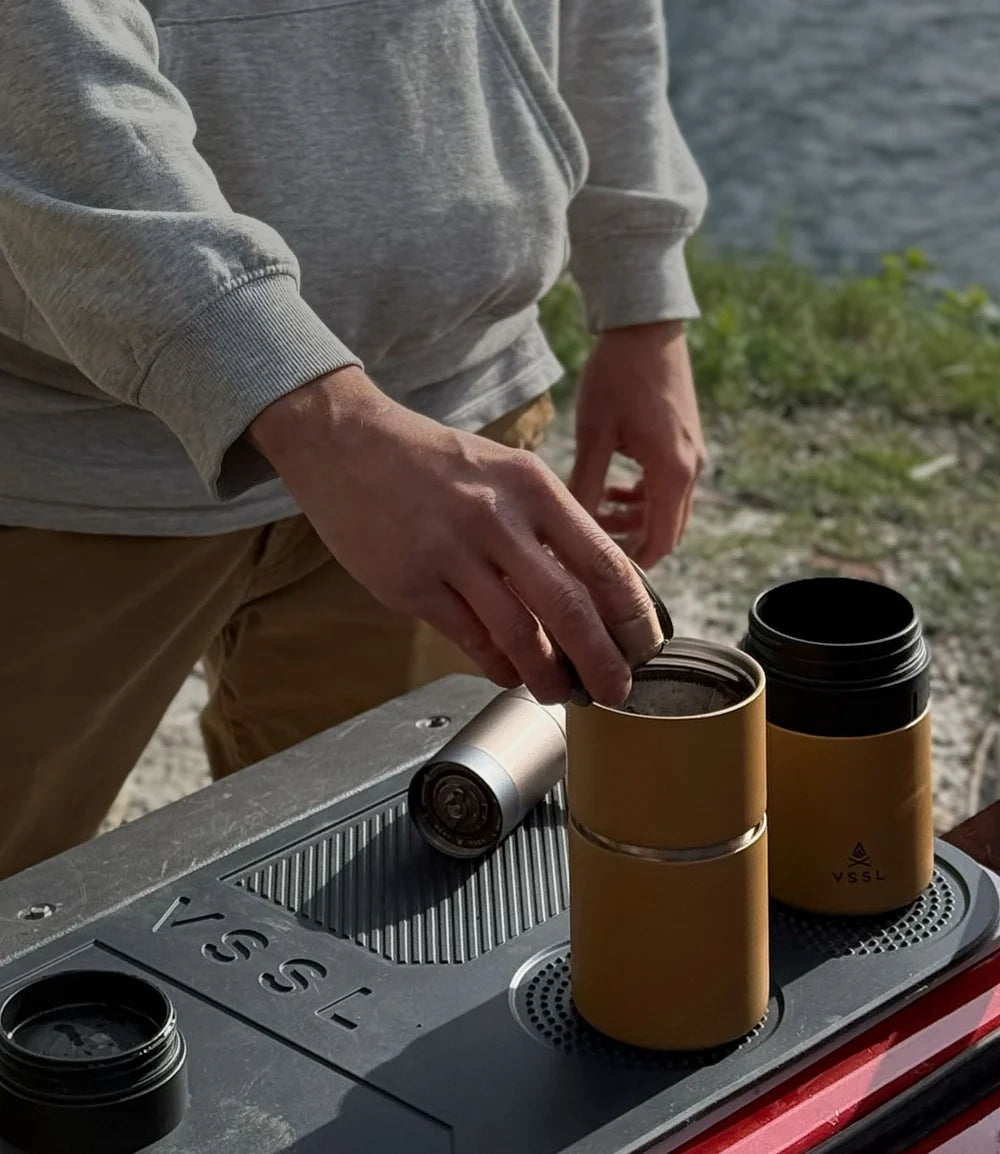 A person outdoors by the water uses a YSSL travel mug to pour coffee into another mug. The YSSL mugs are mustard yellow with black interiors, shown on a black tray.