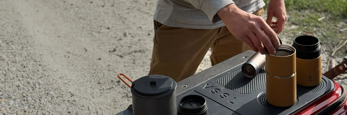 A person outdoors organizes camping cookware on a black folding table, including two amber-brown tumblers, a black lid, and a metal canister.
