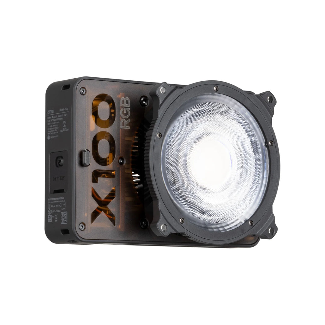 A X100 RGB portable LED light unit with a large circular clear lens on the front, a black side housing, and gold branding, shown on a plain white background.