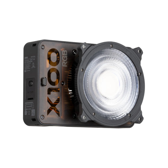 A X100 RGB portable LED light unit with a large circular clear lens on the front, a black side housing, and gold branding, shown on a plain white background.