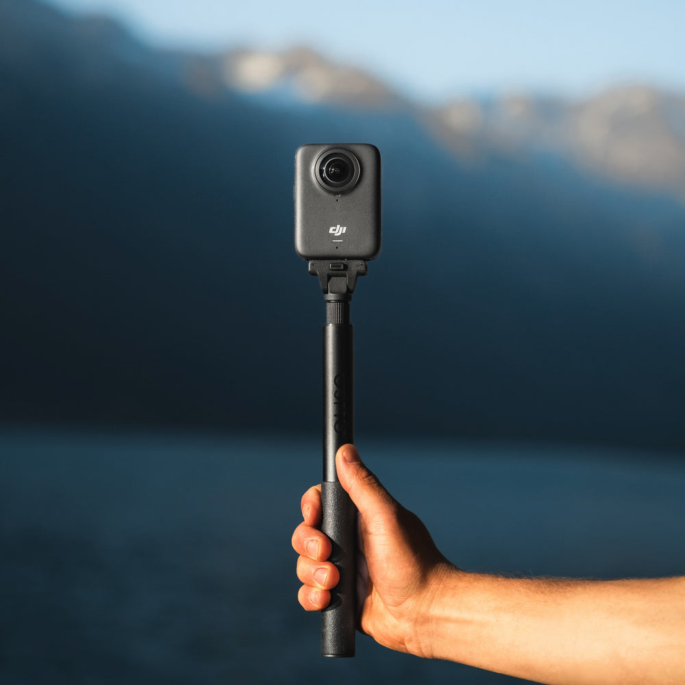 Hand holding a DJI action camera on a pole with a blurred mountain and blue sky background