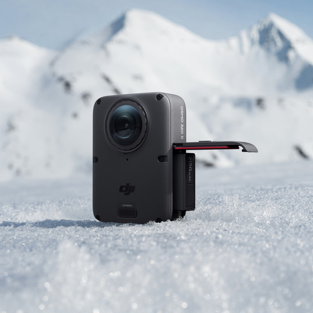 A black DJI action camera with an extended flip-out screen, standing in snow with a blurred mountain range in the background.