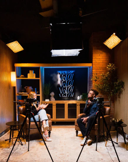 Two people are seated in brown leather chairs, facing each other, in a dimly lit studio with wood-paneled walls. They are being filmed by two cameras on tripods in the foreground, with professional lighting equipment mounted on the ceiling and walls.