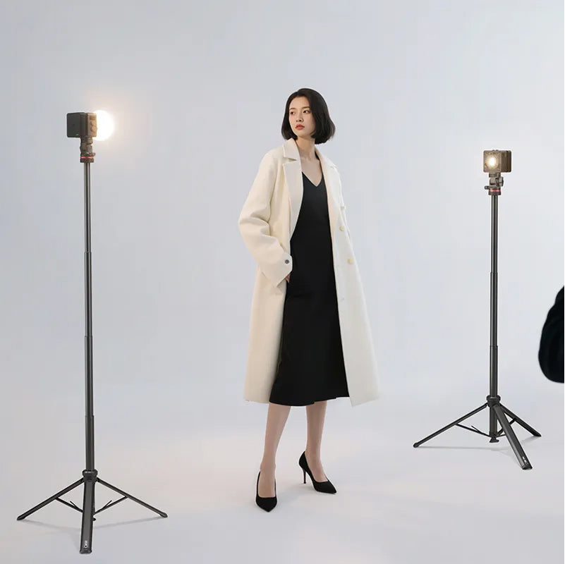 Woman in a white coat and black dress standing between two studio lights on a light gray background