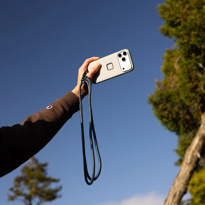 Person holding a phone with a strap against a blue sky with trees