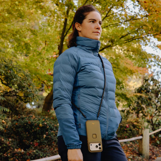 Woman wearing a blue puffer jacket with a phone case attached, standing in an autumnal park.