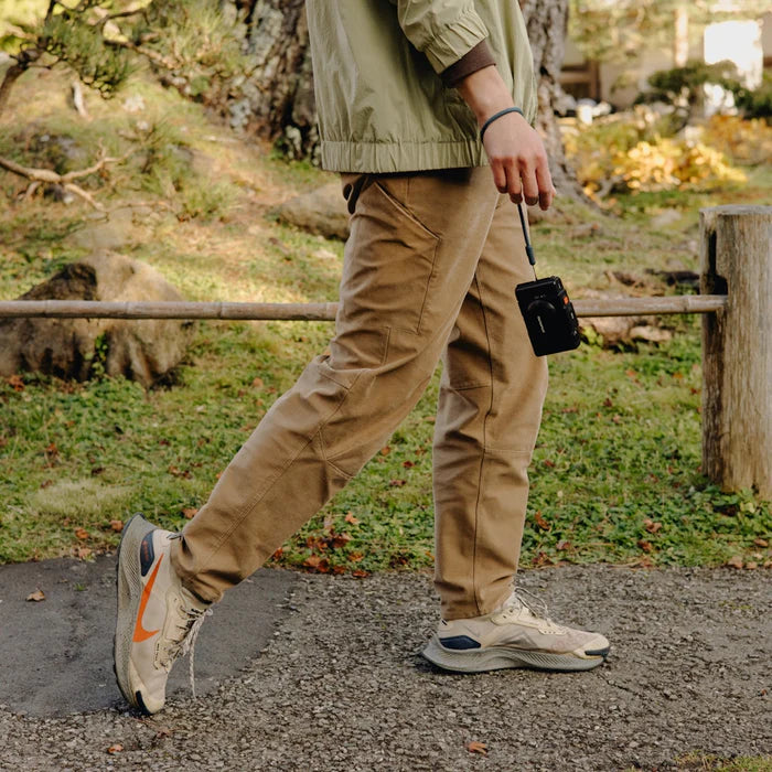 Person walking outdoors wearing beige pants and a light jacket, holding a black device.