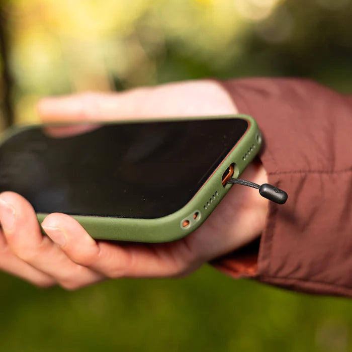 Person holding a green phone case with a black peak design mobile strap adapter against a blurred natural background